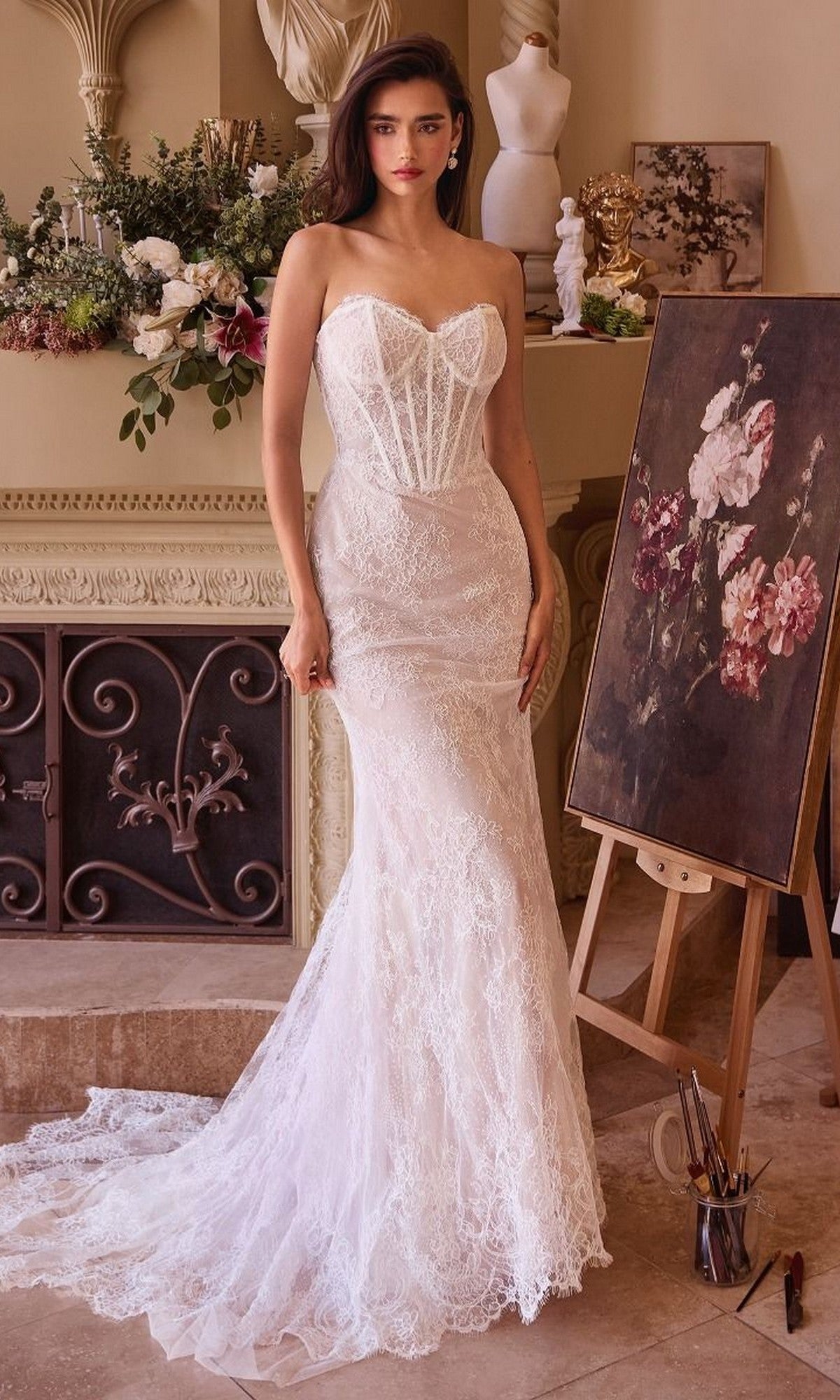 A woman wearing the Andrea and Leo WL044 Long Formal Dress, featuring a strapless silhouette and delicate lace detailing, stands indoors near an easel with a floral painting and a decorated mantle, embodying the elegance of bridal celebrations.